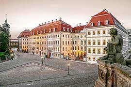 Außenansicht des Taschenbergpalais: lange barocke Fassade mit roten Dächern über dem gepflasterten Platz, rechts im Vordergrund eine Sandsteinfigur