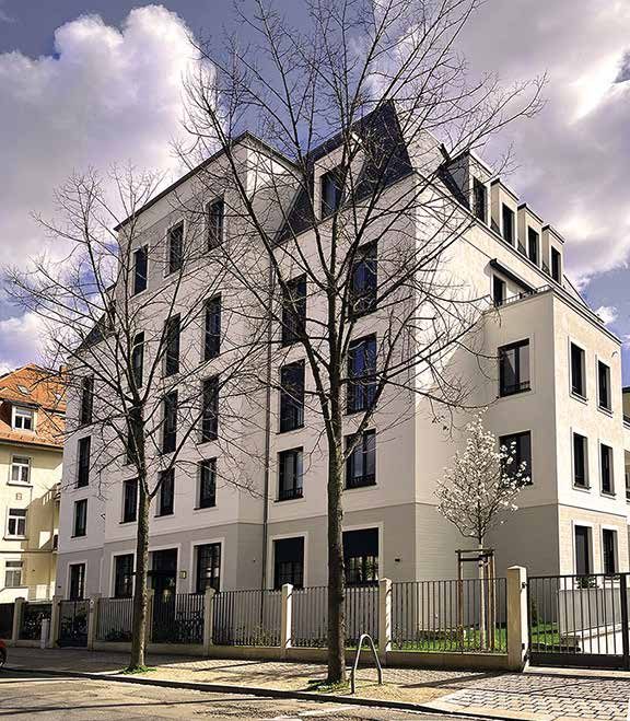 Foto der fertiggestellten Mehrfamilienvilla Therese: weiße Eckfassade mit Mansarddach hinter kahlen Straßenbäumen und Zaun