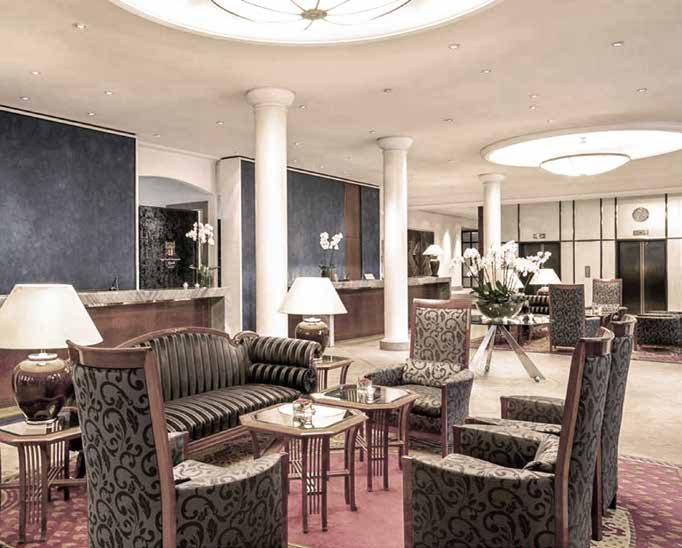 Die Lobby vor der Sanierung: Sitzgruppe mit gestreiftem Sofa und ornamental gemusterten Polstersesseln auf rotem Teppich, Tischlampen, weiße Säulen und dahinter Empfangstresen vor dunkelblauen Wänden