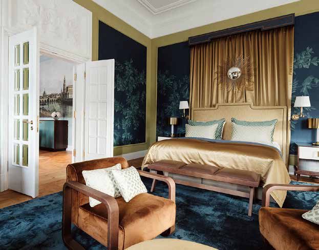Schlafzimmer der Royal Suite: Bett vor goldenem Vorhang mit Sonnenspiegel, dunkelblaue Wandfelder mit Pflanzenmotiven in goldenen Rahmen, cognacfarbene Polstersessel auf blauem Teppich; durch die offene Flügeltür Blick auf die Dresden-Ansicht im Nebenzimmer